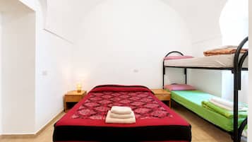 A single bed with a red and black bedspread, a bedside table with a lamp, and a bunk bed to the right.