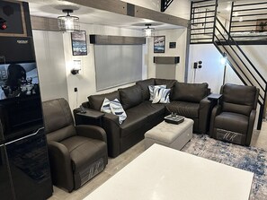 A modern living room with a sectional sofa, a coffee table, and a TV. There is a staircase leading to an upper level. - Cover RV Pad w/Large 45ft Destination RV & Outdoor Bar / Hot Tub (Bolivar Peninsula)