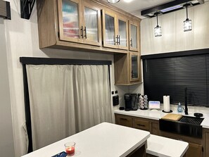A compact kitchen with wooden cabinets, a countertop, a coffee maker, and a black-framed window. - Cover RV Pad w/Large 45ft Destination RV & Outdoor Bar / Hot Tub (Bolivar Peninsula)