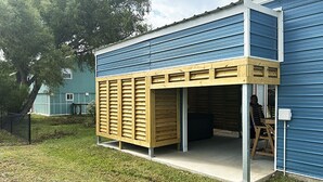 A building with a blue exterior, a wooden structure attached to the side, and a person inside. - Cover RV Pad w/Large 45ft Destination RV & Outdoor Bar / Hot Tub (Bolivar Peninsula)