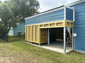 A building with a blue exterior, a wooden structure attached to the side, and a person inside. - Cover RV Pad w/Large 45ft Destination RV & Outdoor Bar / Hot Tub (Bolivar Peninsula)