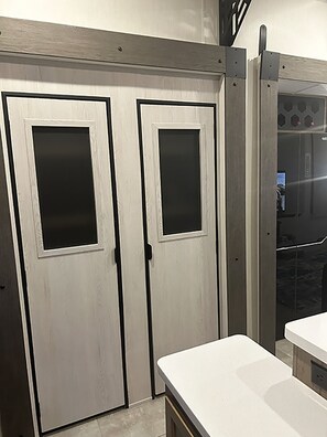 Habitació amb dues portes tancades, una superfície de treball blanca i una vista parcial d'un bany amb dutxa.