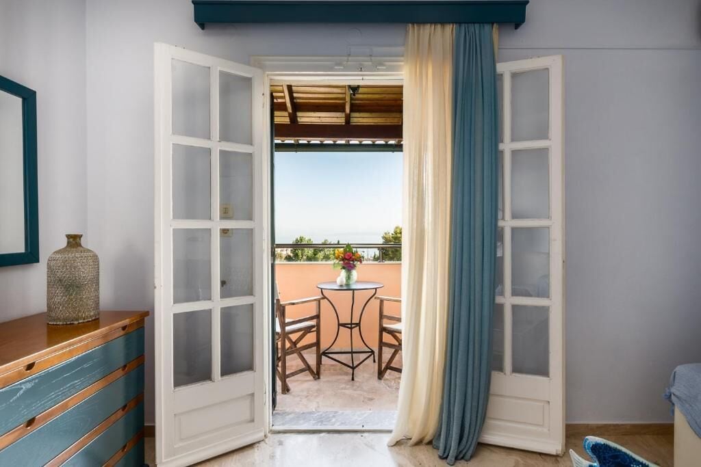 Uma porta aberta que dá para uma varanda com mesa e cadeiras, um vaso com flores e um aparador de madeira.