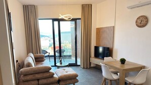 A modern living room with a sofa, ottoman, dining table with chairs, a TV, and a view through large glass doors. - Sunny Stay apartment in Ksamil (Ksamil)