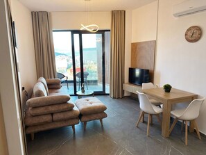 Ein modernes Wohnzimmer mit Sofa, Hocker, Esstisch mit Stühlen, einem Fernseher und Blick durch große Glasüren.