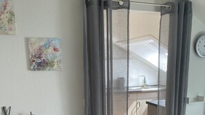 A modern room with a wooden table, a dark armchair, a kitchen area, and a window with curtains. - Studio Apartment Braun (Wasserburg am Bodensee)