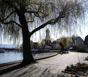 A park with a large tree, a body of water, and buildings in the background. - Studio Apartment Braun (Wasserburg am Bodensee)