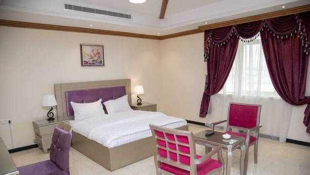 A hotel room with a bed, a desk, two chairs, a nightstand, a lamp, and a window with curtains. - Diva Serviced Apartments (Jeddah)