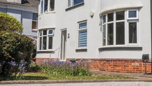 Une maison à deux étages, avec une base en briques et des murs supérieurs blancs, dotée de nombreuses fenêtres et d’un petit jardin agrémenté de fleurs violettes.