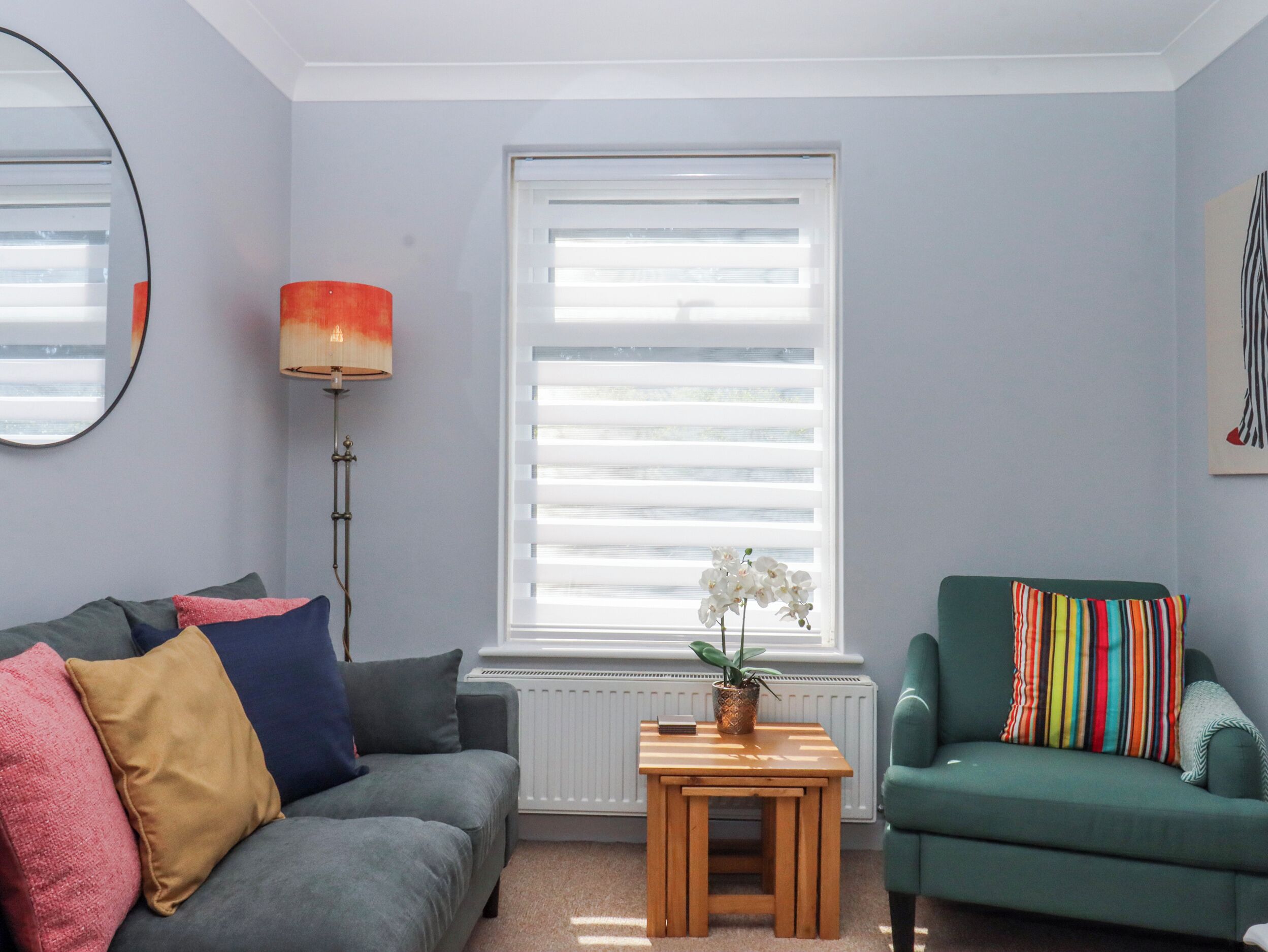 A living room with a grey sofa, a green armchair, a small wooden table with a plant, and a radiator.