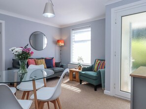 A modern living room with a glass dining table, a sofa, a green armchair, a round mirror, and a floor lamp. - The Arches (Torquay)