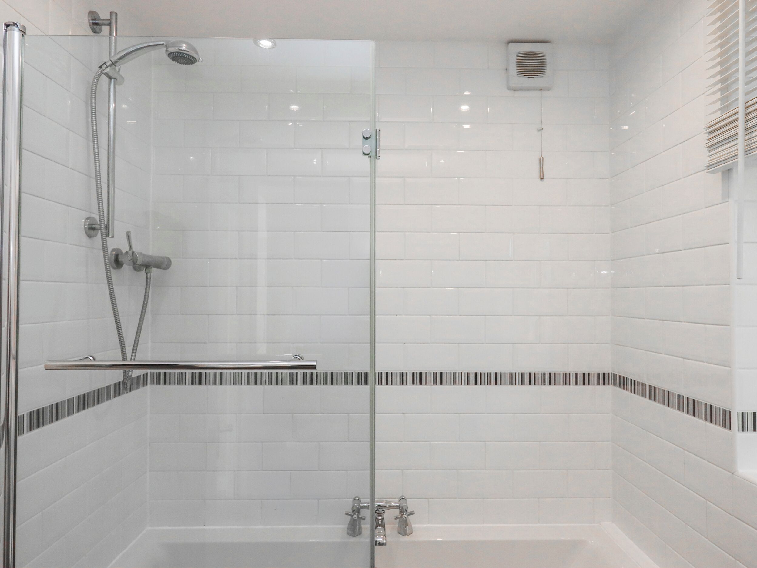 Un espace douche clos par des parois de verre, avec une baignoire, des murs recouverts de carreaux blancs et une bordure de carreaux à rayures.