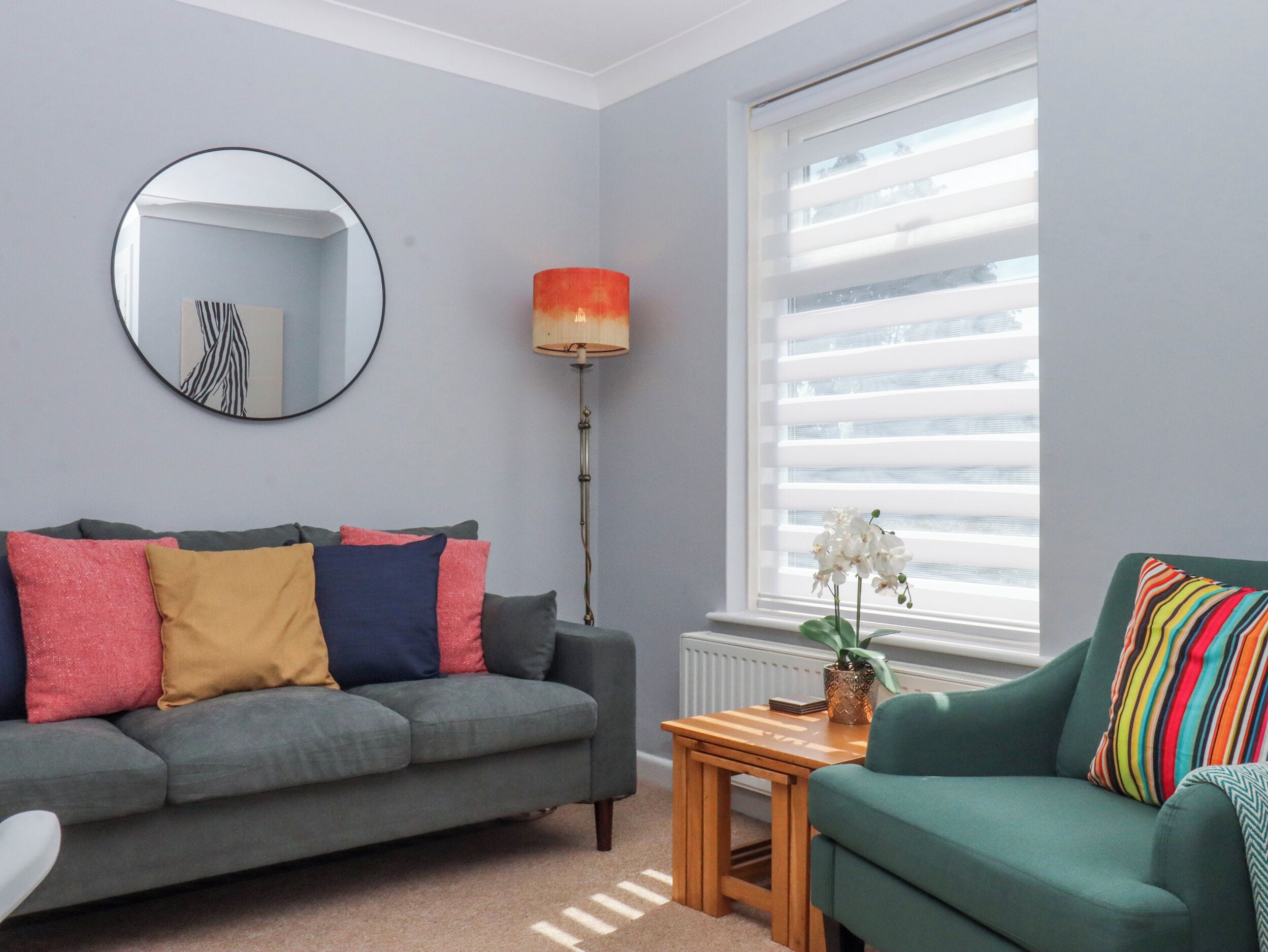 Un salon avec un canapé gris, un fauteuil vert, un miroir rond, une lampe sur pied et une fenêtre avec des stores.