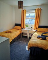 A hotel room with two beds, a desk, a potted plant, and a window with curtains.