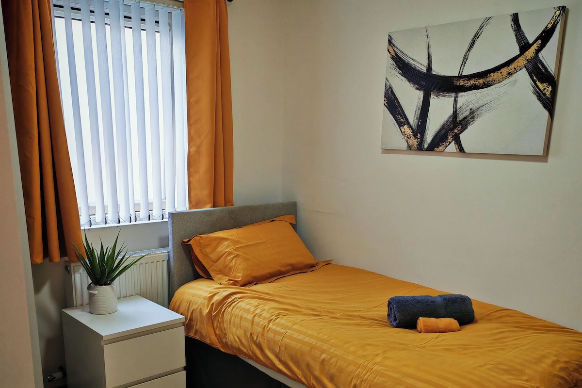 Un dormitorio con cama, mesita de noche, radiador, ventana con cortinas, una obra de arte en la pared y un tapete marrón.