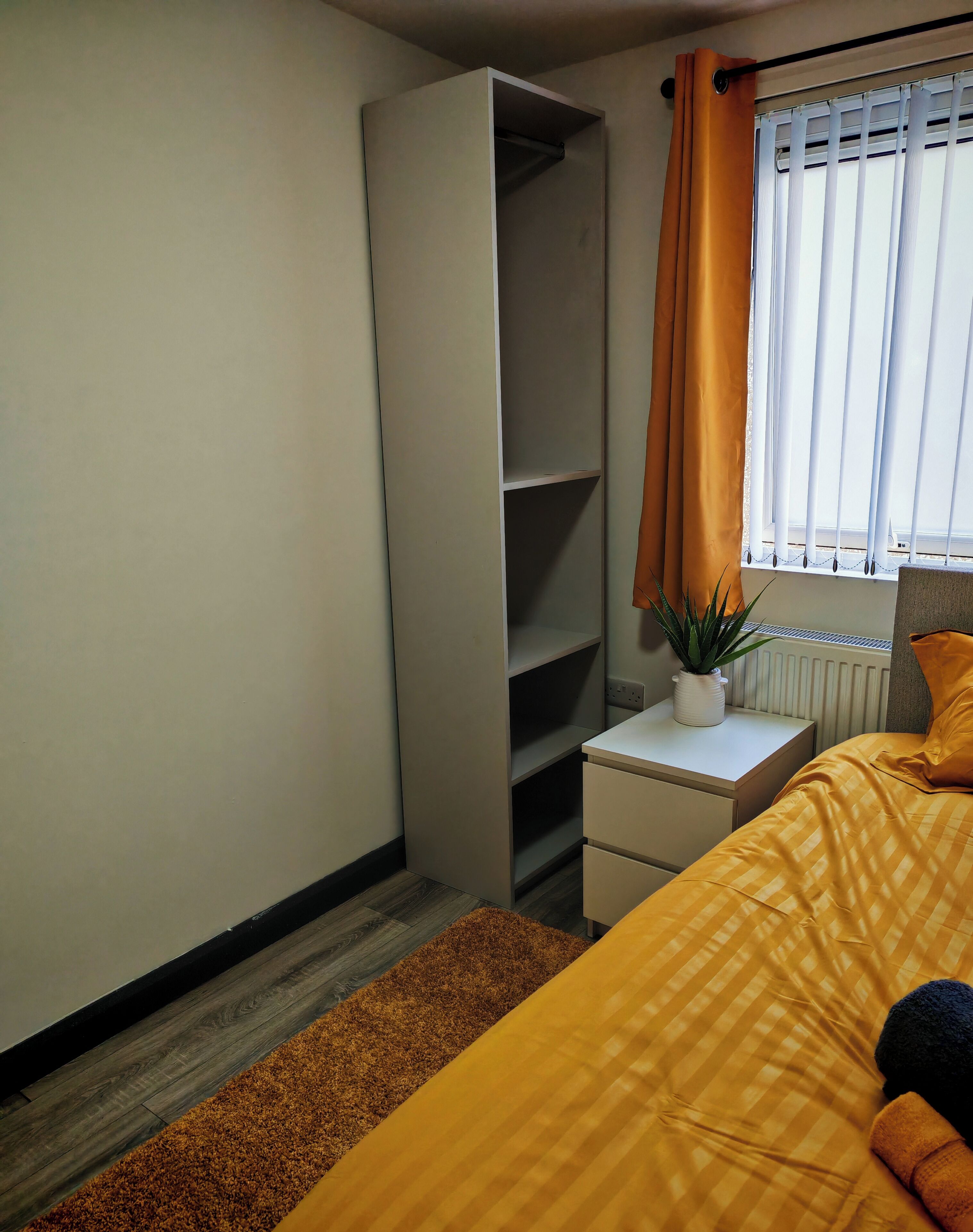 A bedroom with a bed, a nightstand, a wardrobe, a radiator, and a window with curtains.