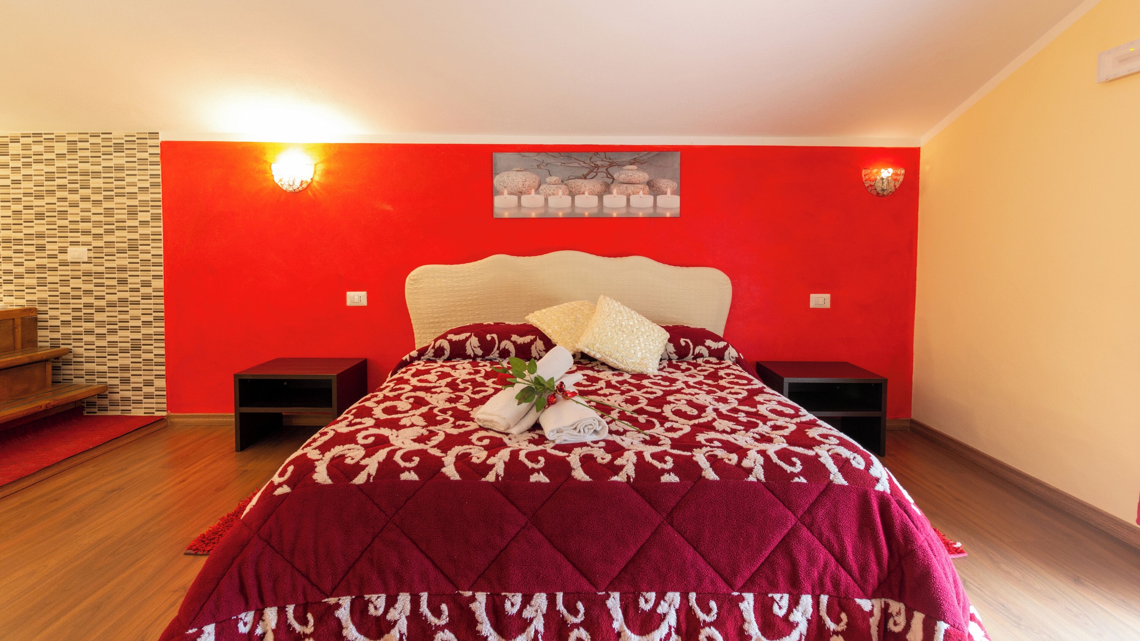 A bedroom with a red wall, a bed with a patterned bedspread, two bedside tables, and a wall-mounted light fixture.