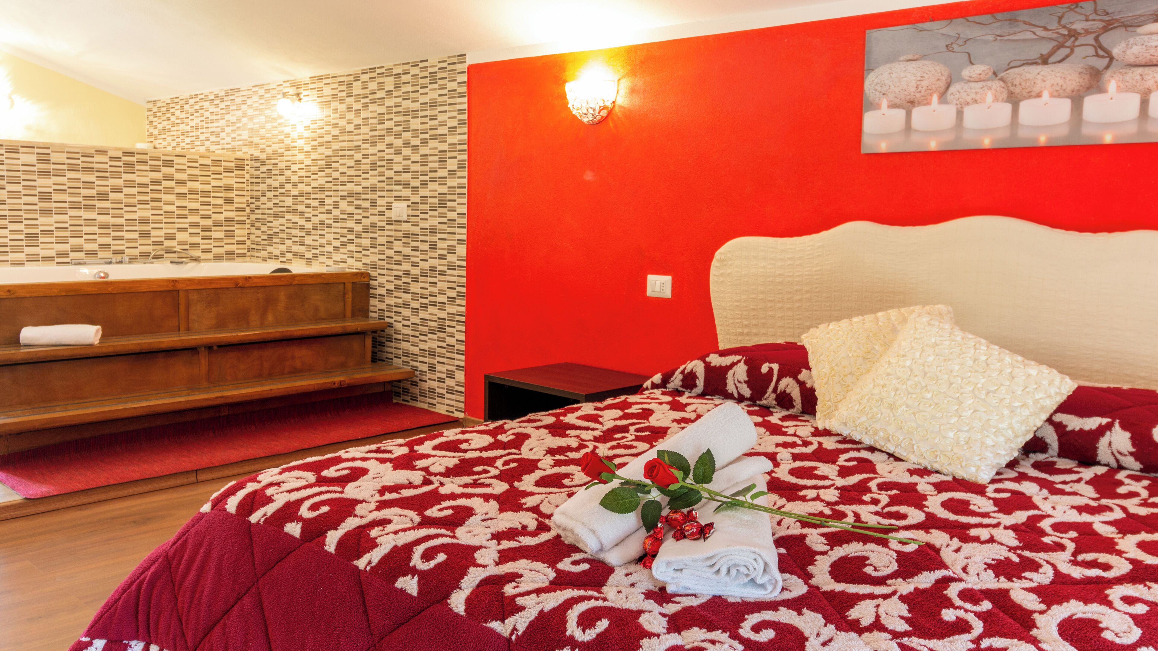 Une chambre d’hôtel avec un mur rouge, un banc en bois, un lit recouvert d’une courtepointe à motifs rouges et blancs, et un tableau représentant des pierres.