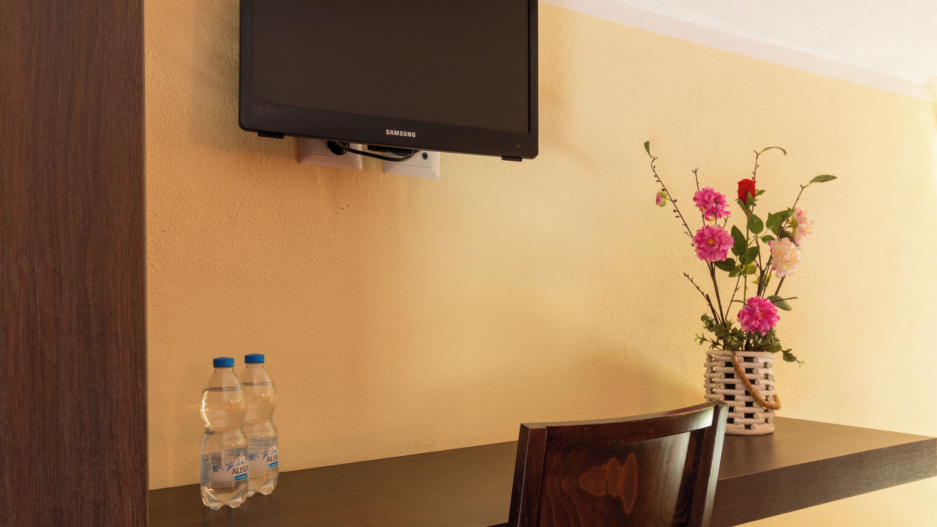 A hotel room with a flat-screen TV mounted on the wall, a wooden table with two water bottles, and a vase with pink flowers.