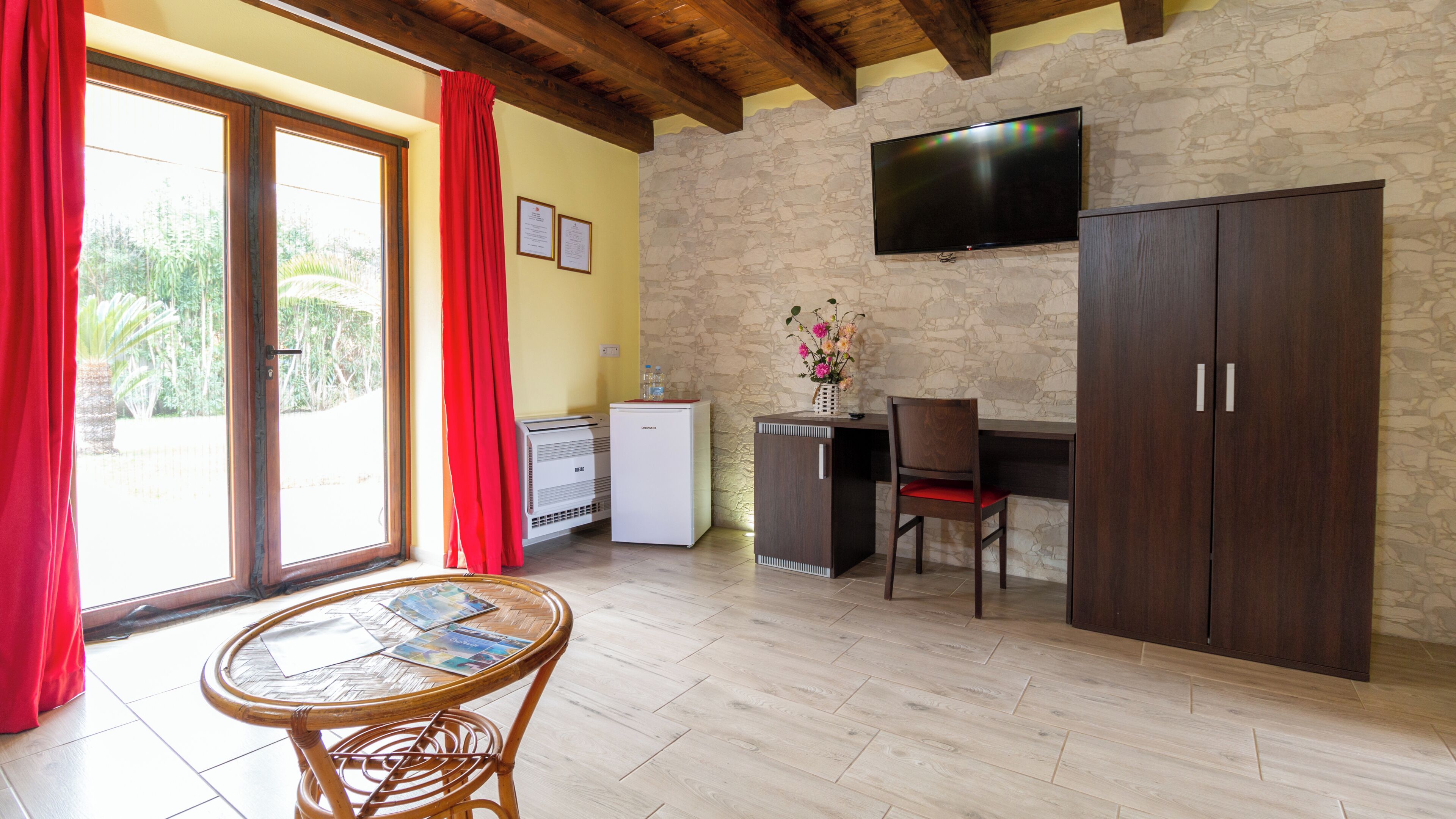 A room with a stone wall, a flat-screen TV, a wooden desk, a chair, a small refrigerator, a table with a woven chair, and a window with red curtains.