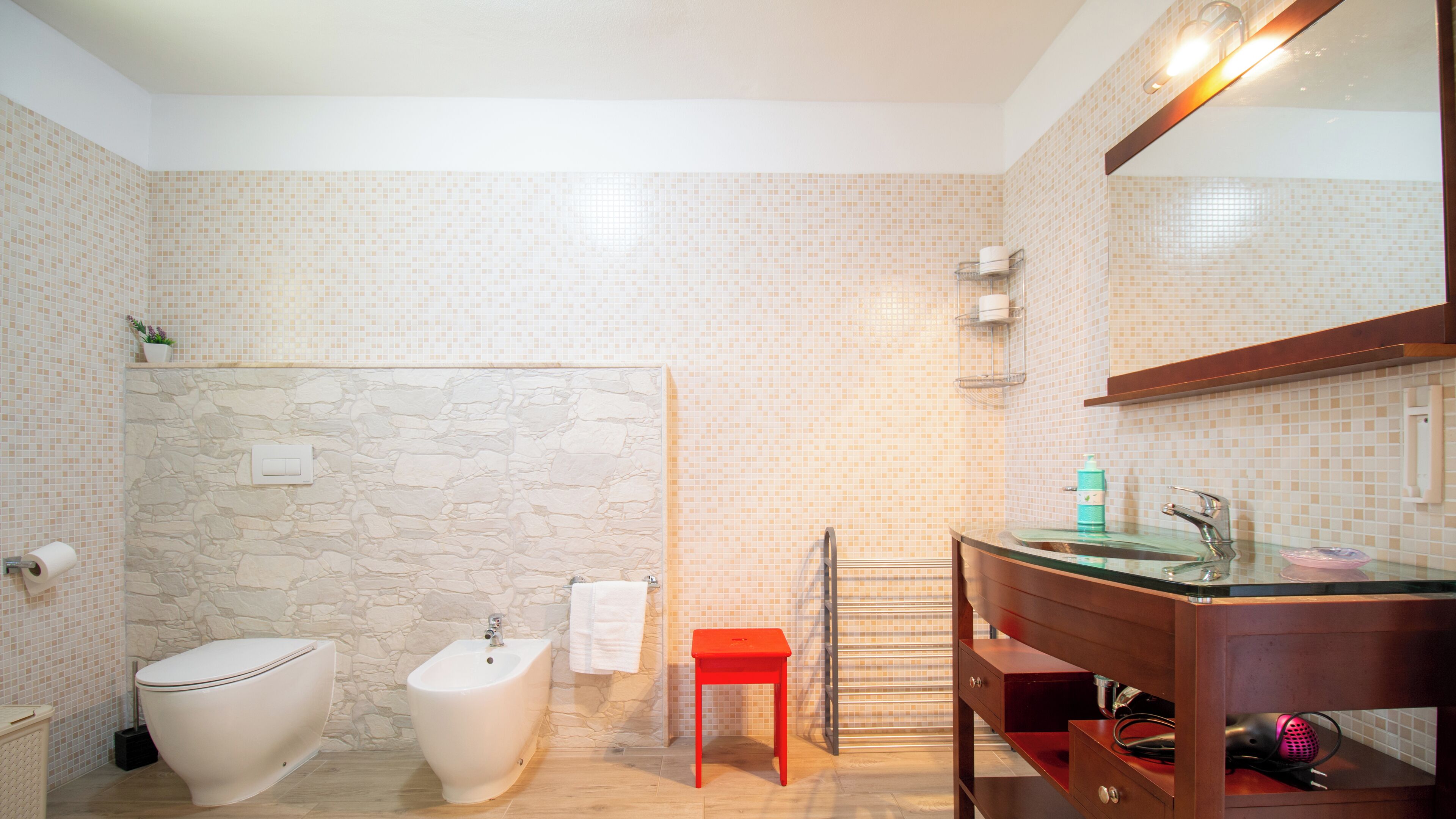 A bathroom with a stone wall, a bidet, a toilet, a red stool, a glass sink, and a towel rack.