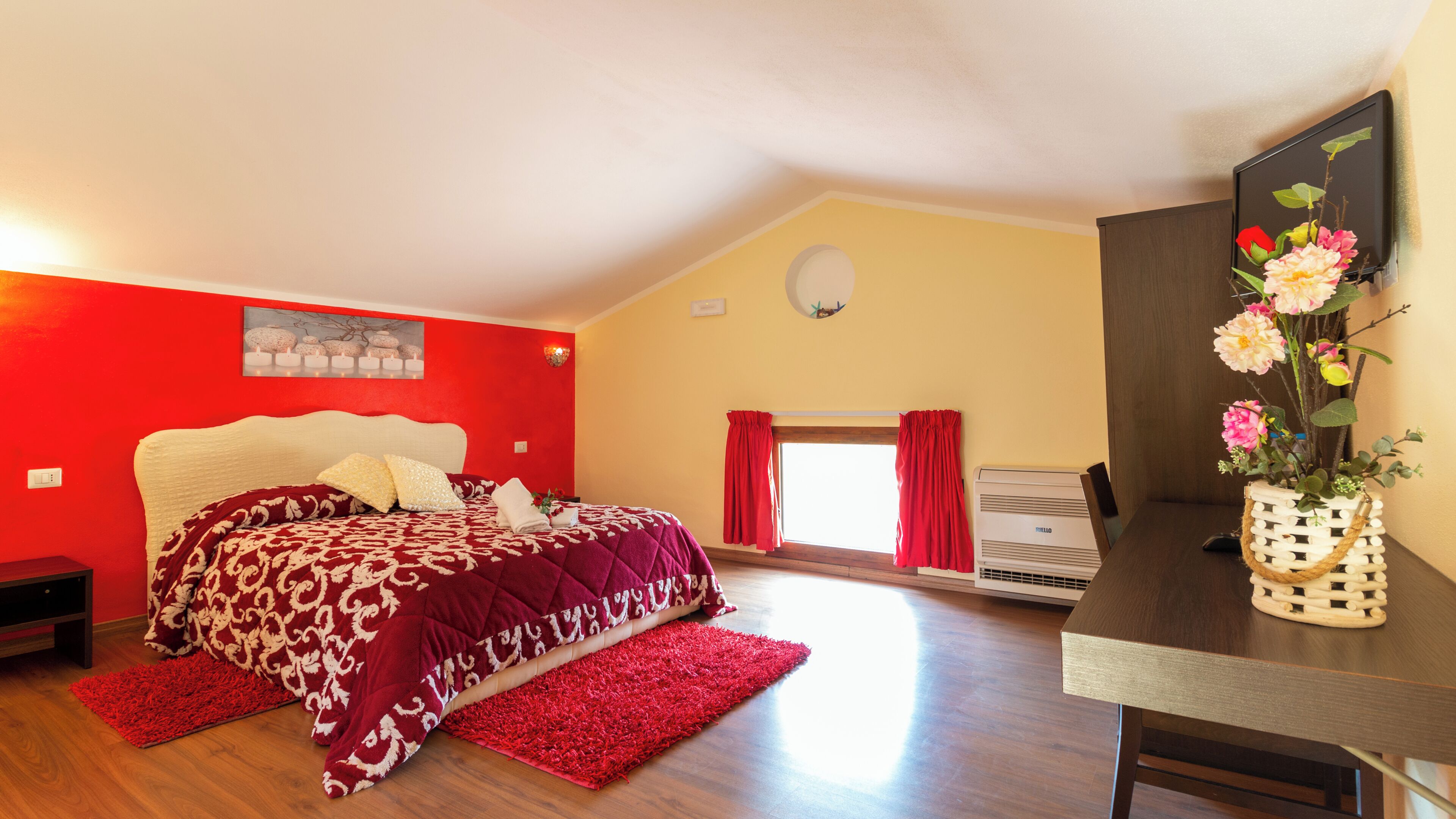 A bedroom with a bed, a red rug, a desk with a vase of flowers, and a window with curtains.