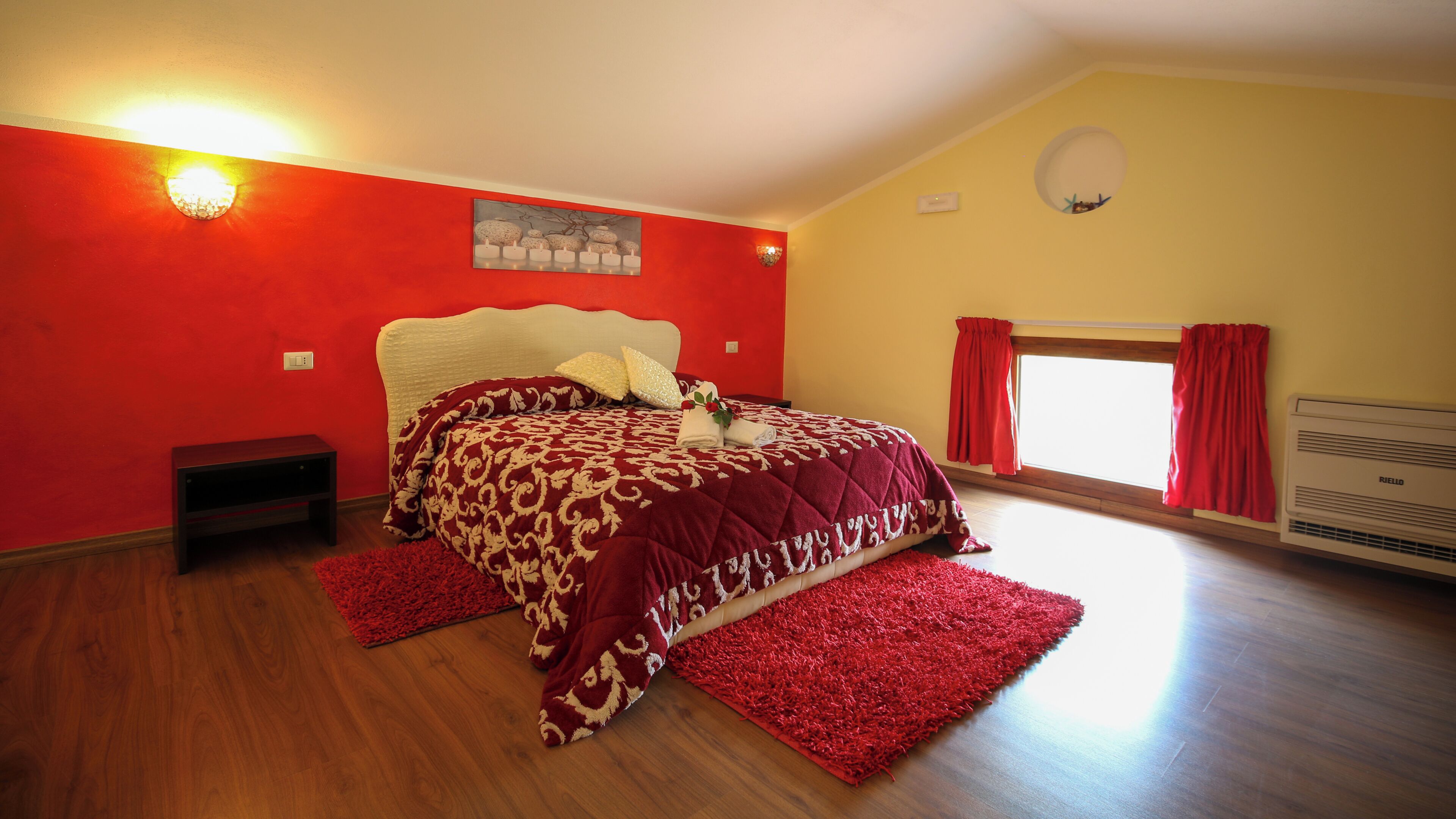 A bedroom with a large bed, red walls, a painting, a window with curtains, and a radiator.