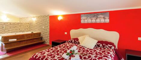 A bedroom with a red wall, a large bed with a white headboard, a wooden bench, a red and white patterned bedspread, and a bouquet of flowers.