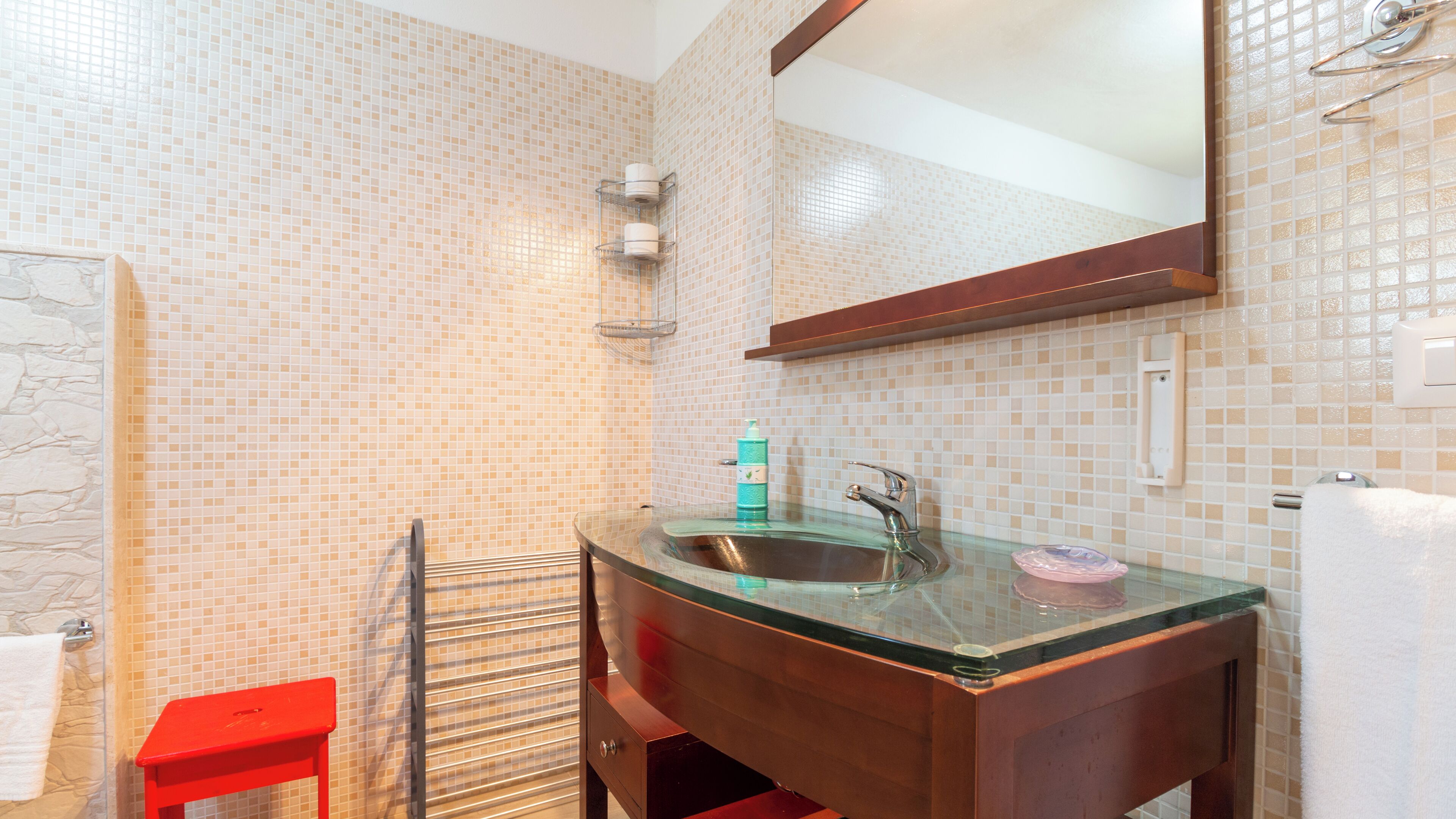 A bathroom with a glass sink, a wooden vanity, a red stool, and a tiled wall.