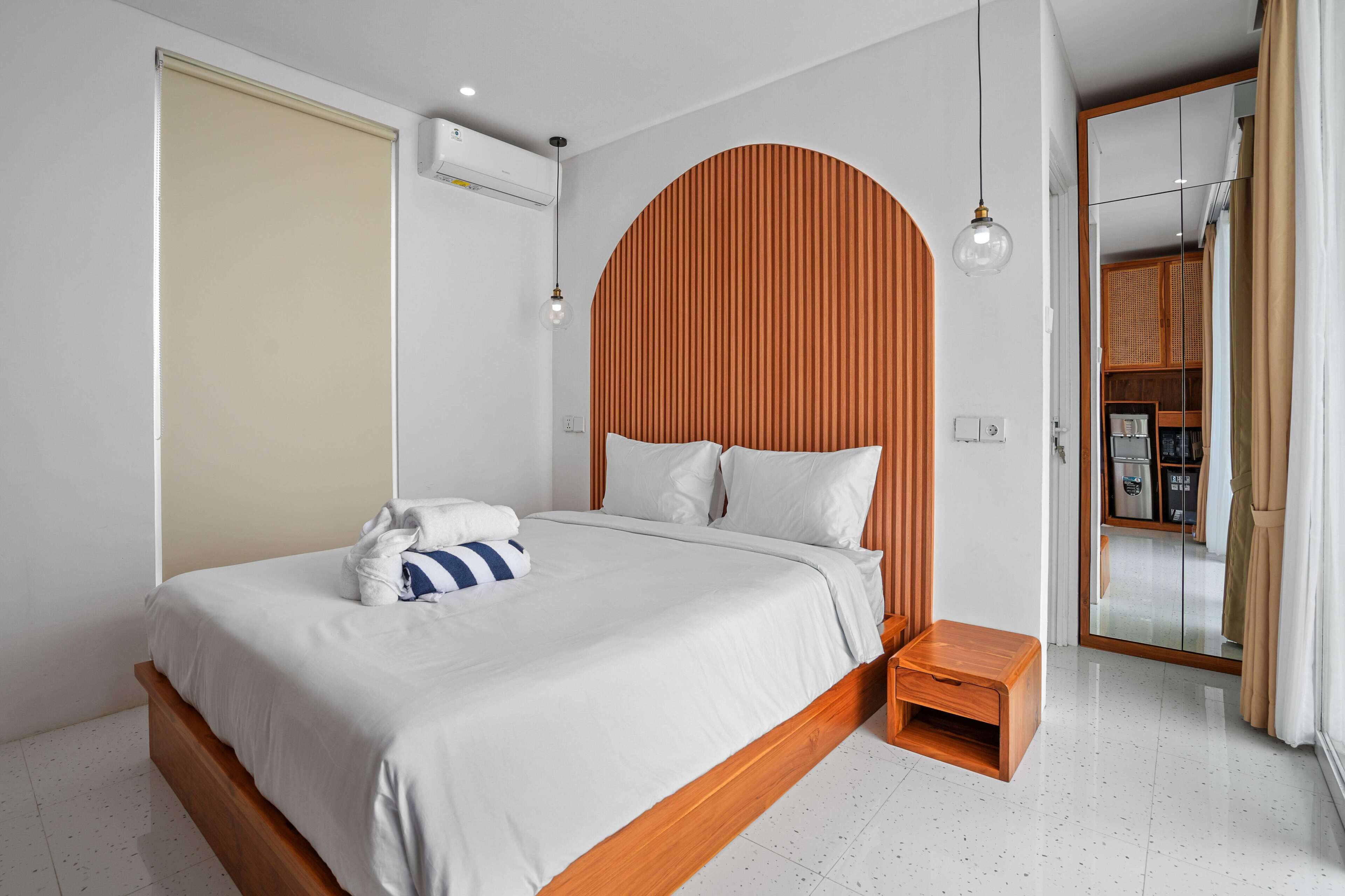 A modern bedroom with a wooden headboard, a bed with white bedding, a small wooden nightstand, and a mirror with a wooden frame.