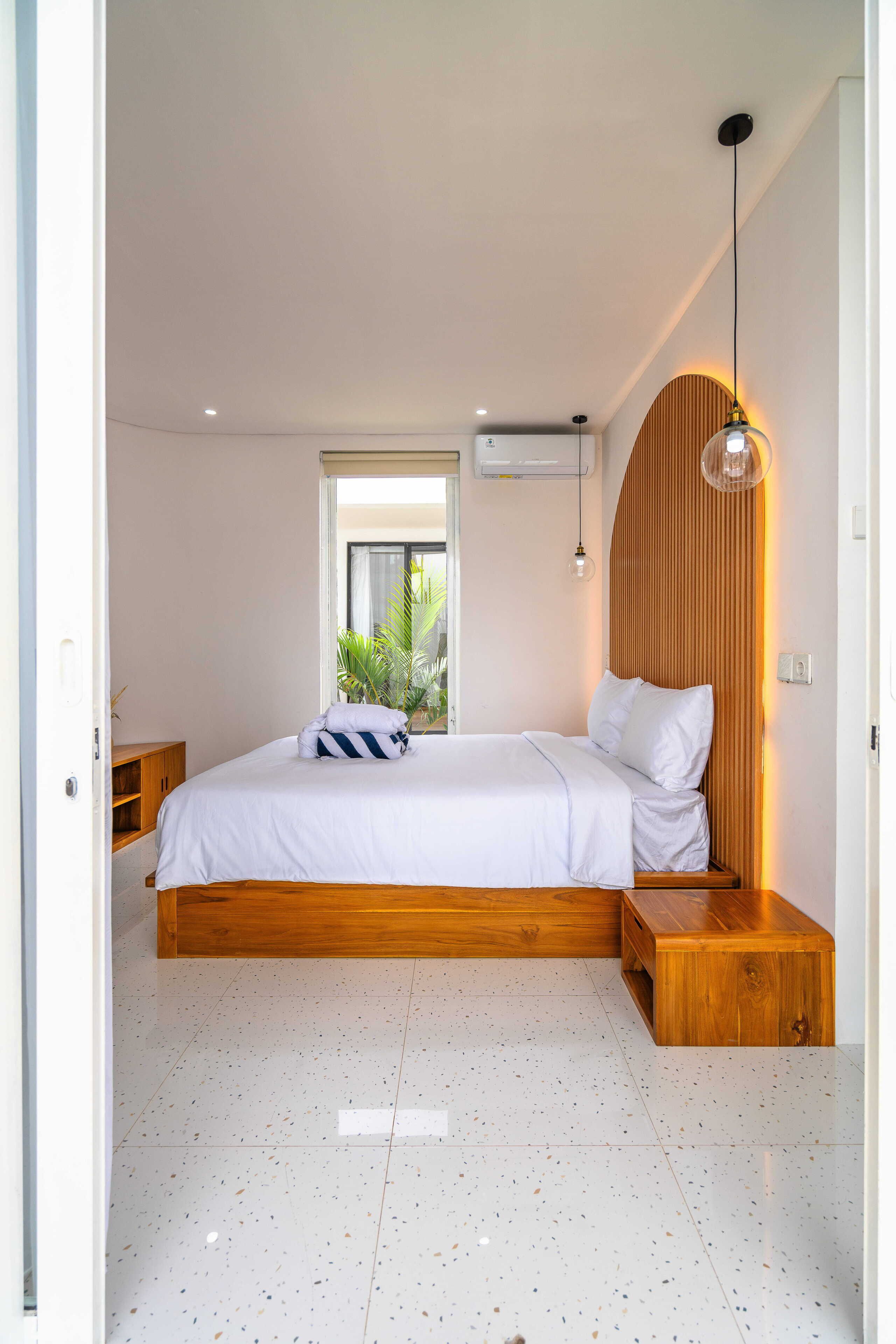 A modern bedroom with a wooden bed frame, a bedside table, a hanging light fixture, and a window with a view of greenery.