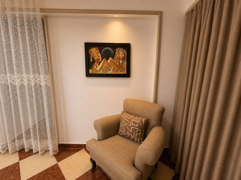 Ein Zimmer mit einem beigefarbenen Sessel, einem gemusterten Kissen, einem an der Wand hängenden Kunstwerk mit Pyramiden, einem gemusterten Vorhang und einem Schachbrettfußboden.