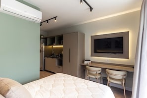 Ein modernes Hotelzimmer mit einem Bett, einem Schreibtisch mit zwei Stühlen, einem an der Wand montierten Fernseher und einer Küchenzeile im Hintergrund.