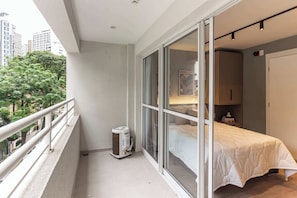 Ein Balkon mit weißer Brüstung, ein Fenster mit Blick auf Bäume und Gebäude, ein Bett mit weißer Bettdecke und ein Heizkörper am Boden.
