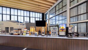 A modern hotel reception area with a large window, a flat-screen TV, and various amenities like a juice dispenser, coffee machine, and food display.