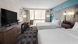 A hotel room with a TV, two beds, a desk with a chair, and a large window with curtains.