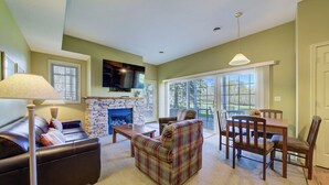 A living room with a fireplace, a sofa, a plaid armchair, a dining table with chairs, and a TV mounted above the fireplace. - Fairway to Heaven II at Shanty Creek (Bellaire)