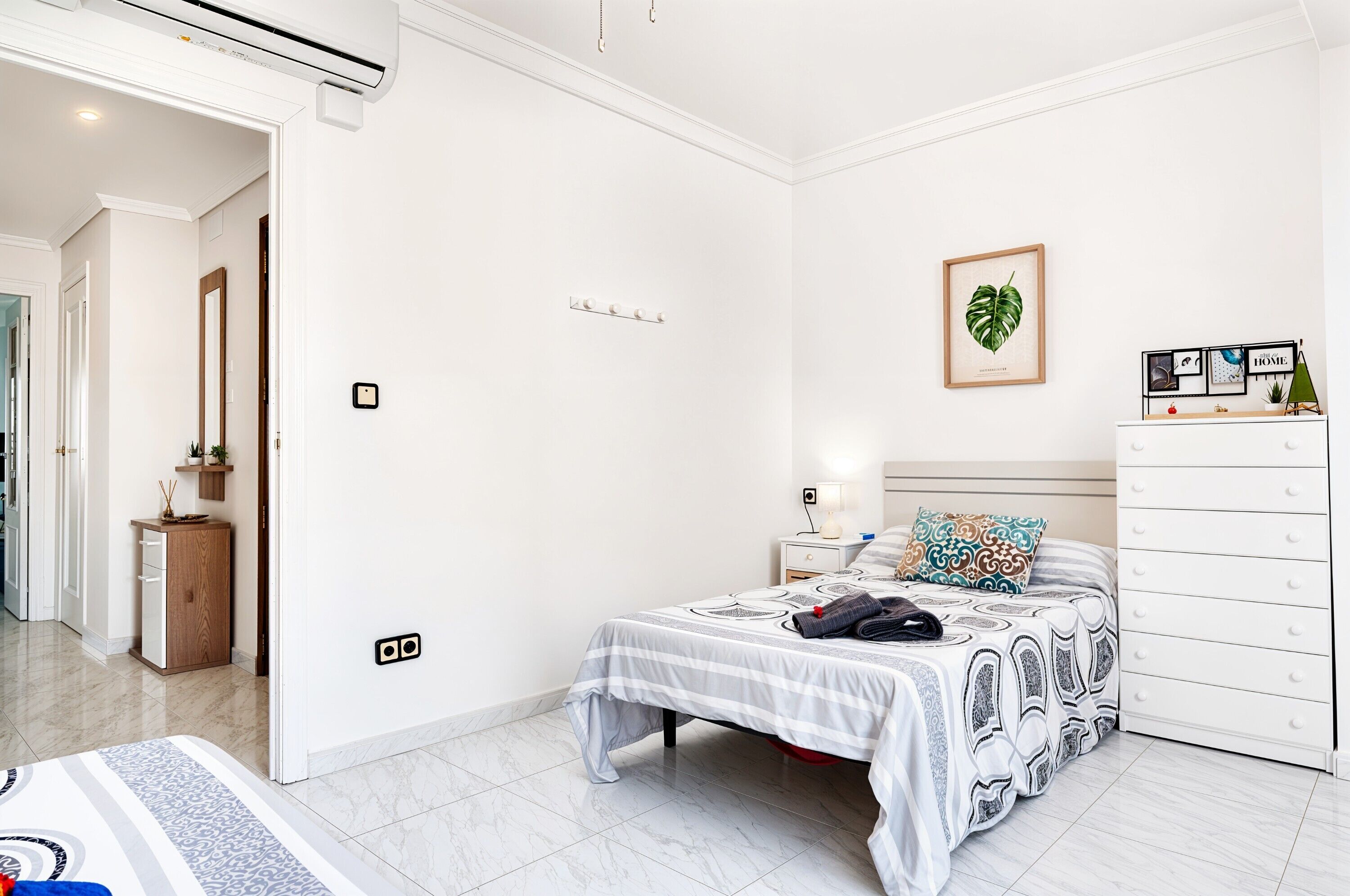 A bedroom with a bed, bedside table, dresser, and a wall-mounted shelf with framed pictures.
