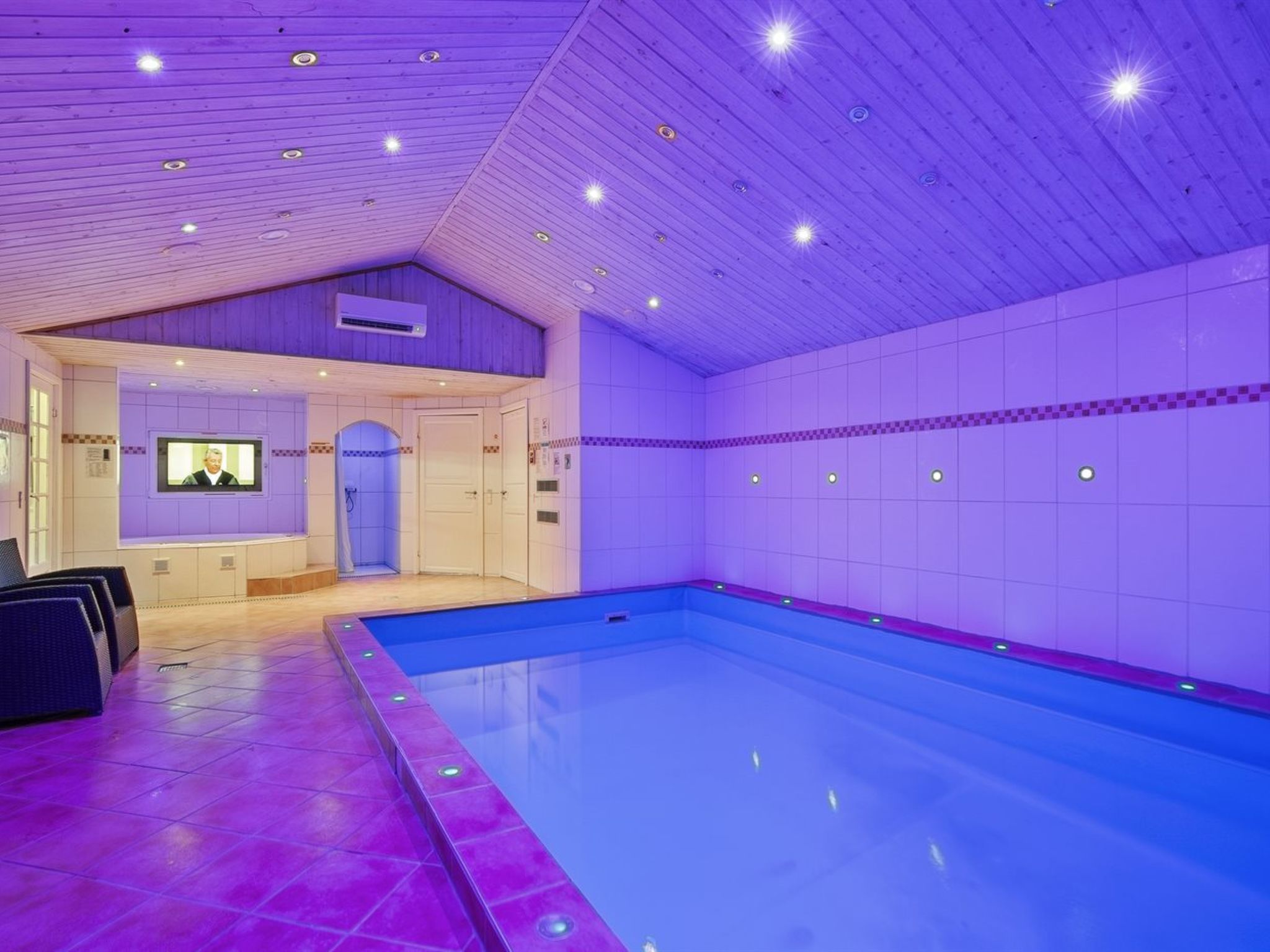 Ein großes Hallenbad mit gefliesten Wänden und Decke, einem Fernsehbildschirm und einer Sitzecke mit Stühlen.