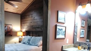 Un dormitorio con techo de madera, una cama con manta escocesa, una mesita de noche con lámpara y un baño con lavamanos y espejo.