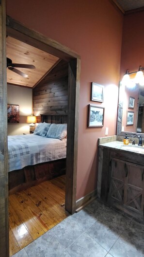 Een slaapkamer met een houten plafond, een bed met een geruite plaid, een nachtkastje met een lamp, en een badkamer met een wastafel en spiegel.