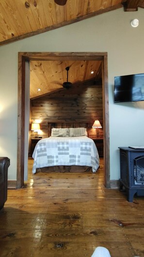 Une chambre avec un plafond en bois, un lit recouvert d’une courtepointe, deux lampes de chevet, un téléviseur fixé au mur et un poêle à bois.