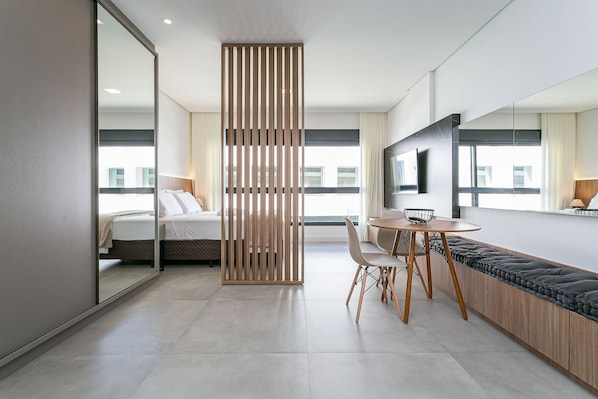 A modern hotel room with a bed, a dining area, a large mirror, and a view of the city. - Modern Studio | Pool | Gym #SG10 (Florianópolis)