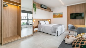 A modern bedroom with a bed, bedside table, chair, TV, and a large window. - Modern Studio | Pool | Gym #SG10 (Florianópolis)