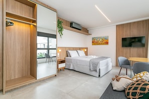 A modern bedroom with a bed, bedside table, chair, TV, and a large window. - Modern Studio | Pool | Gym #SG10 (Florianópolis)