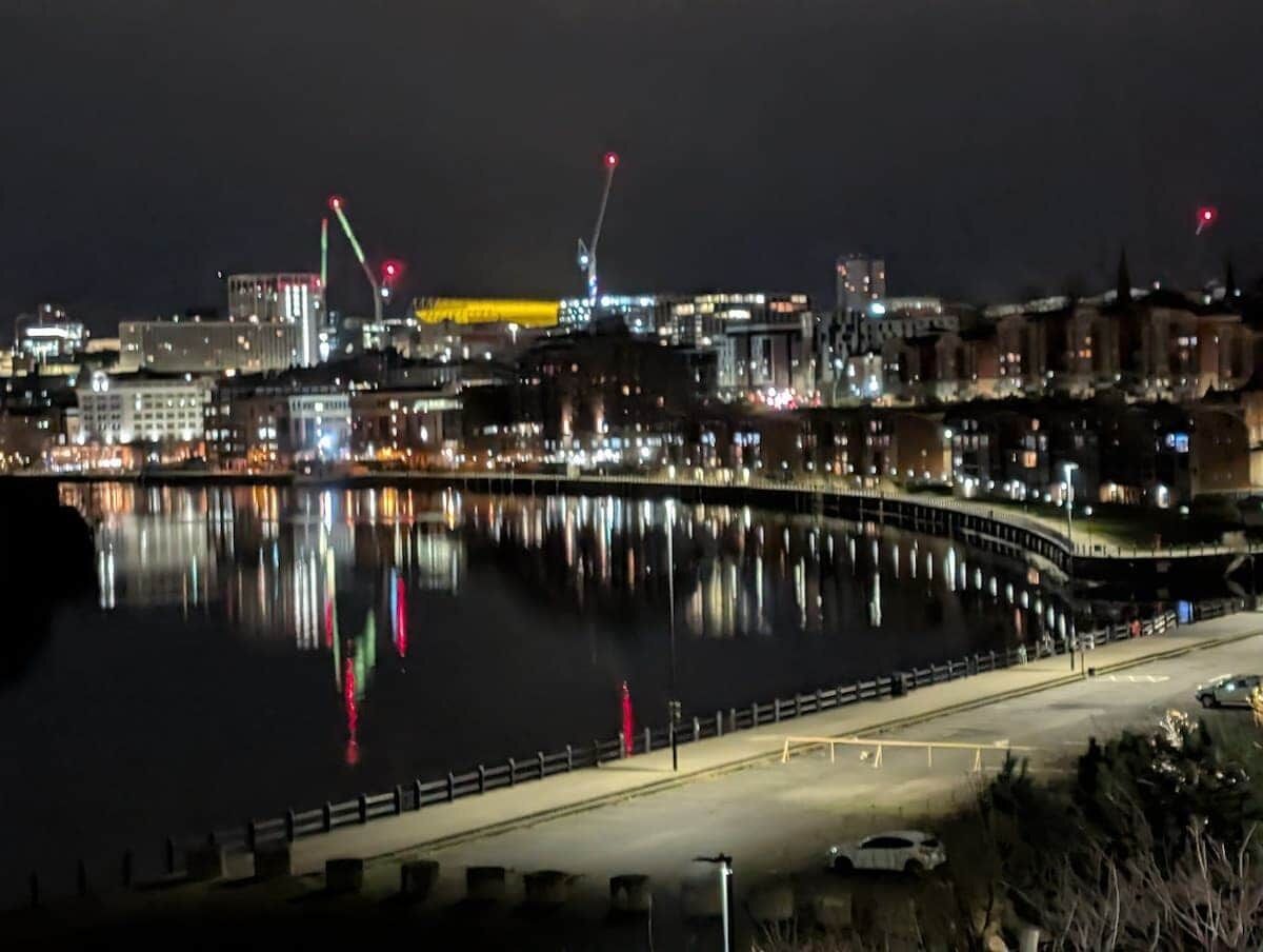 A nighttime cityscape with a waterfront, illuminated buildings, and a bridge with red lights.