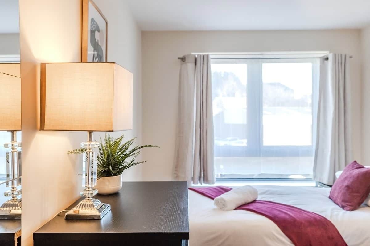 Une chambre d’hôtel avec un lit, un bureau doté d’une lampe, une plante en pot et une fenêtre avec des rideaux.