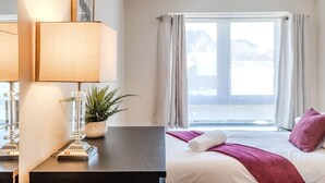 Hotellihuone, jossa on sänky, työpöytä lampun kanssa, ruukkukasvi ja ikkuna, jossa on verhot.