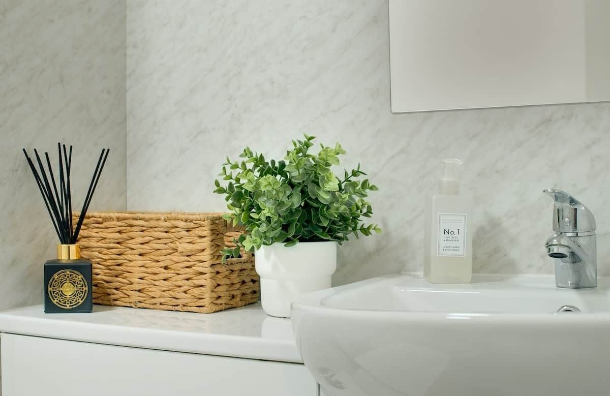 Un baño con pared de mármol, lavamanos blanco, cesta tejida, planta en maceta, difusor de caña y una botella de jabón de manos.
