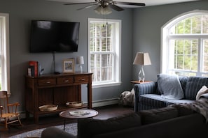 A living room with a television, a sofa, a wooden side table, a lamp, and a window.